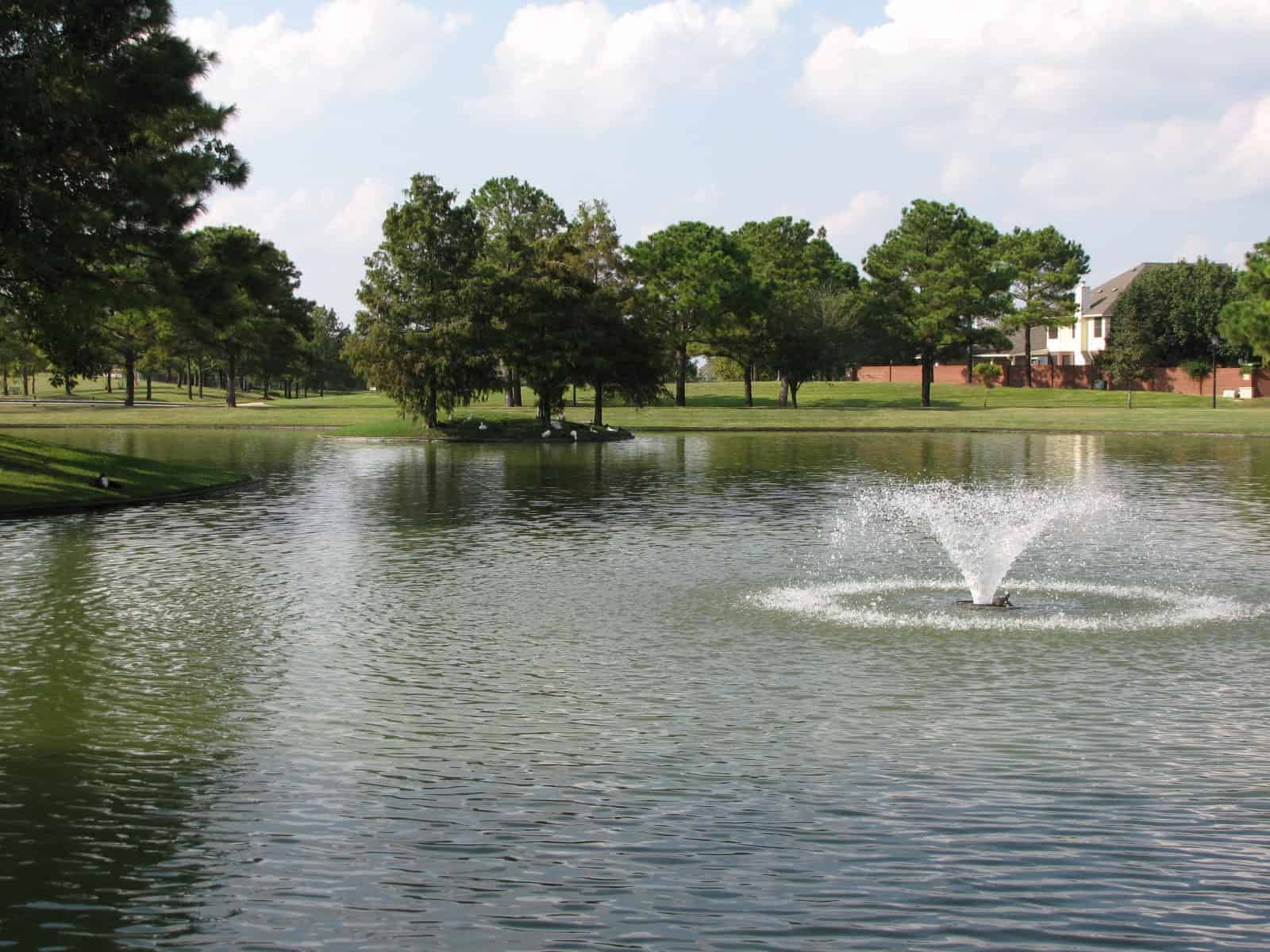 A Copper Lakes lake with a central fountain, a grassy shoreline walking path, and homes on the far bank in Houston, TX 77095