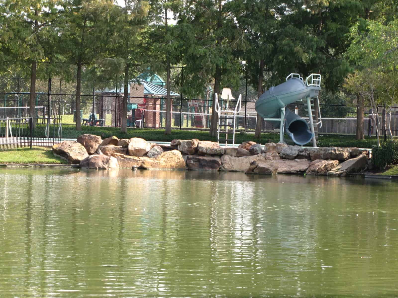 Copper Lakes Realtor โ a community lake with a rocky, grassy shoreline path and homes in Copper Lakes, Houston, TX 77095