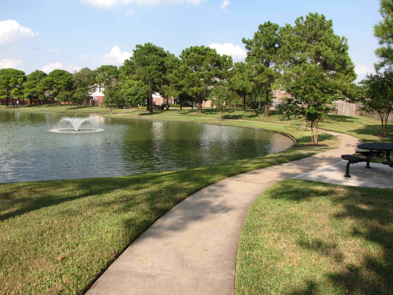 A curved concrete walking path along a grassy lawn beside a Copper Lakes pond with a fountain in Houston, TX 77095