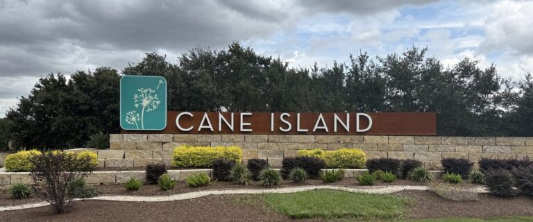 Cane Island stone-and-steel entry monument with a teal dandelion logo panel and landscaped beds, Katy TX 77493