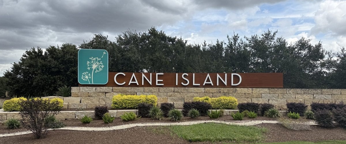 Cane Island stone-and-steel entry monument with a teal dandelion logo panel and landscaped beds, Katy TX 77493