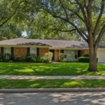 A single-story 1960s brick ranch house on a tree-lined Houston street, the kind of slab-on-grade home that often has cast iron pipes under the slab