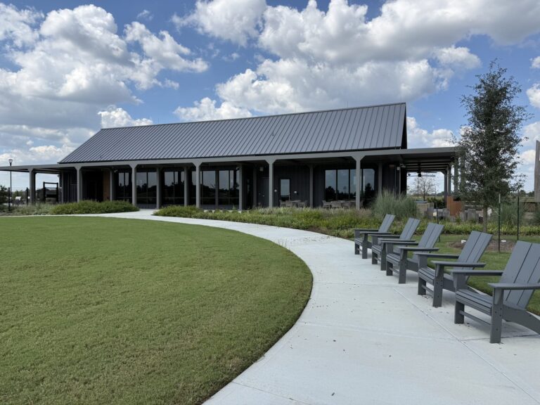 Single-story Everly amenity center with a dark metal roof and a curved walkway across the lawn, Hockley TX 77447