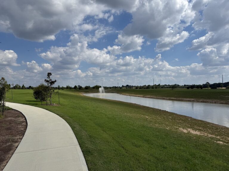 Concrete walking trail curving beside a pond with a fountain in Everly, Hockley TX 77447