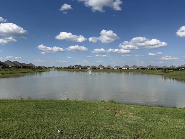 Wide view of the Oakwood Estates lake with a fountain and a row of new homes along the far shore, Waller TX 77484