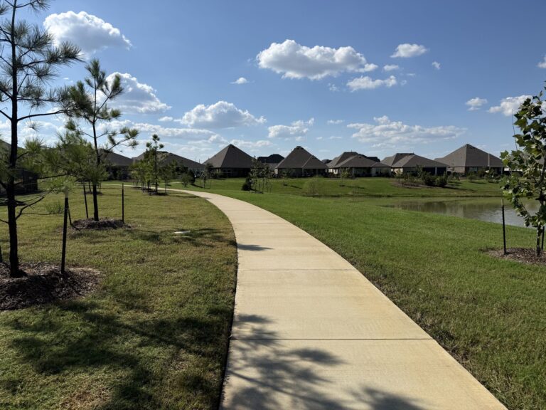 Curving concrete walking trail through young trees beside a pond, with homes in the distance, Oakwood Estates, Waller TX 77484