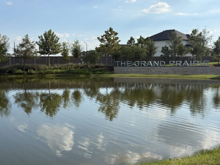 THE GRAND PRAIRIE letters on a stone wall across a pond, with trees and a two-story home behind, Hockley TX 77447