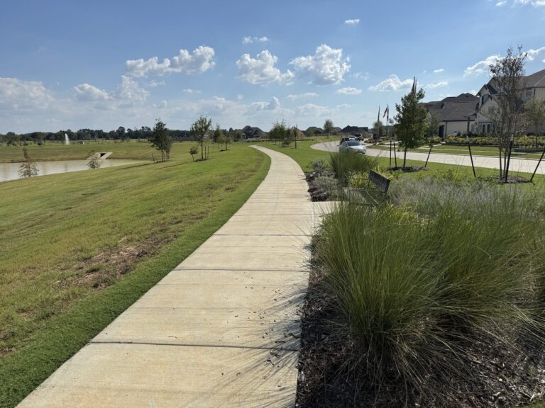 Concrete walking trail curving along a pond with ornamental grasses and new homes behind it, Wildrye, Waller TX 77484